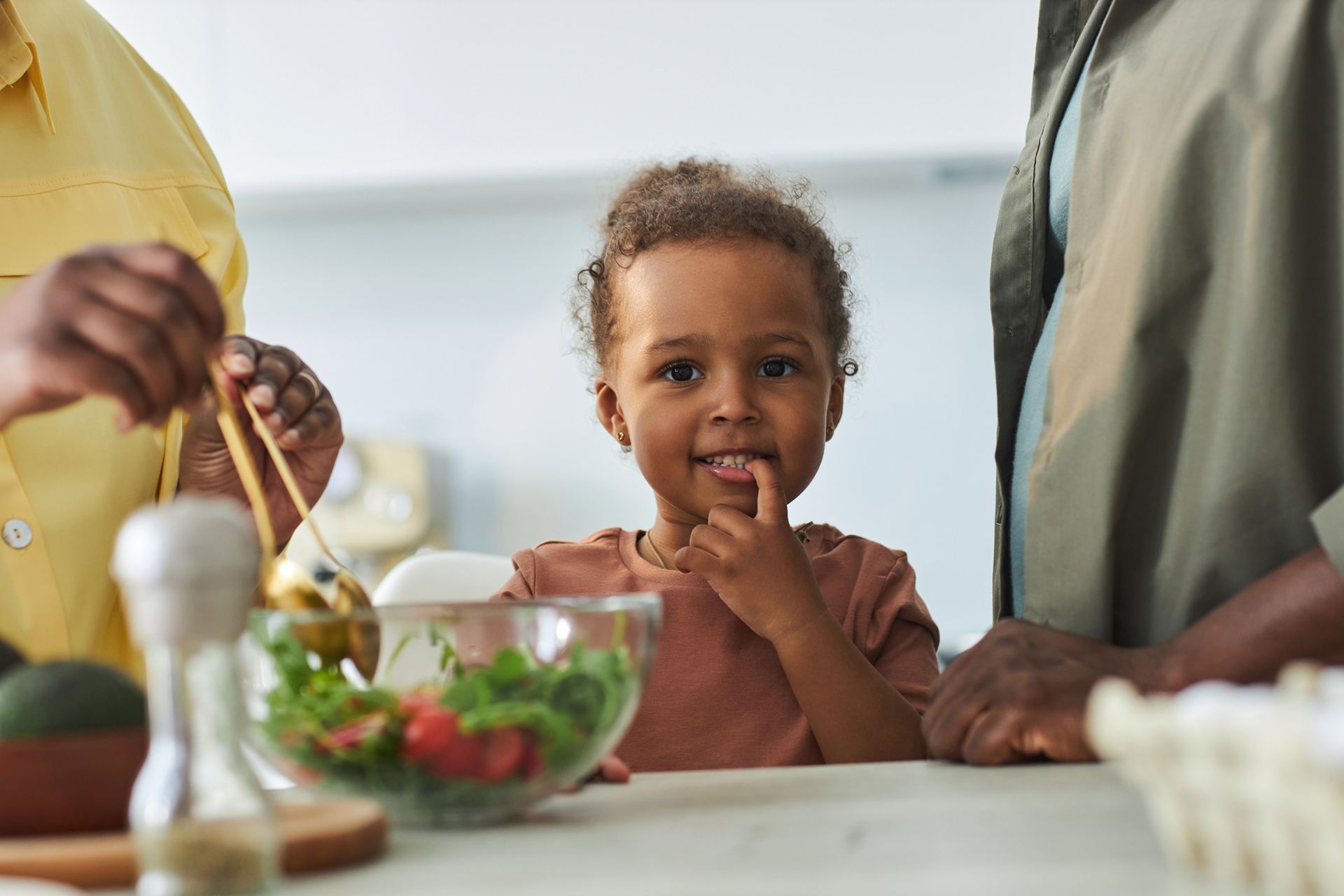 Little girl puts a finger to her lips and smiles while adults around her prepare a salad in the kitchen.