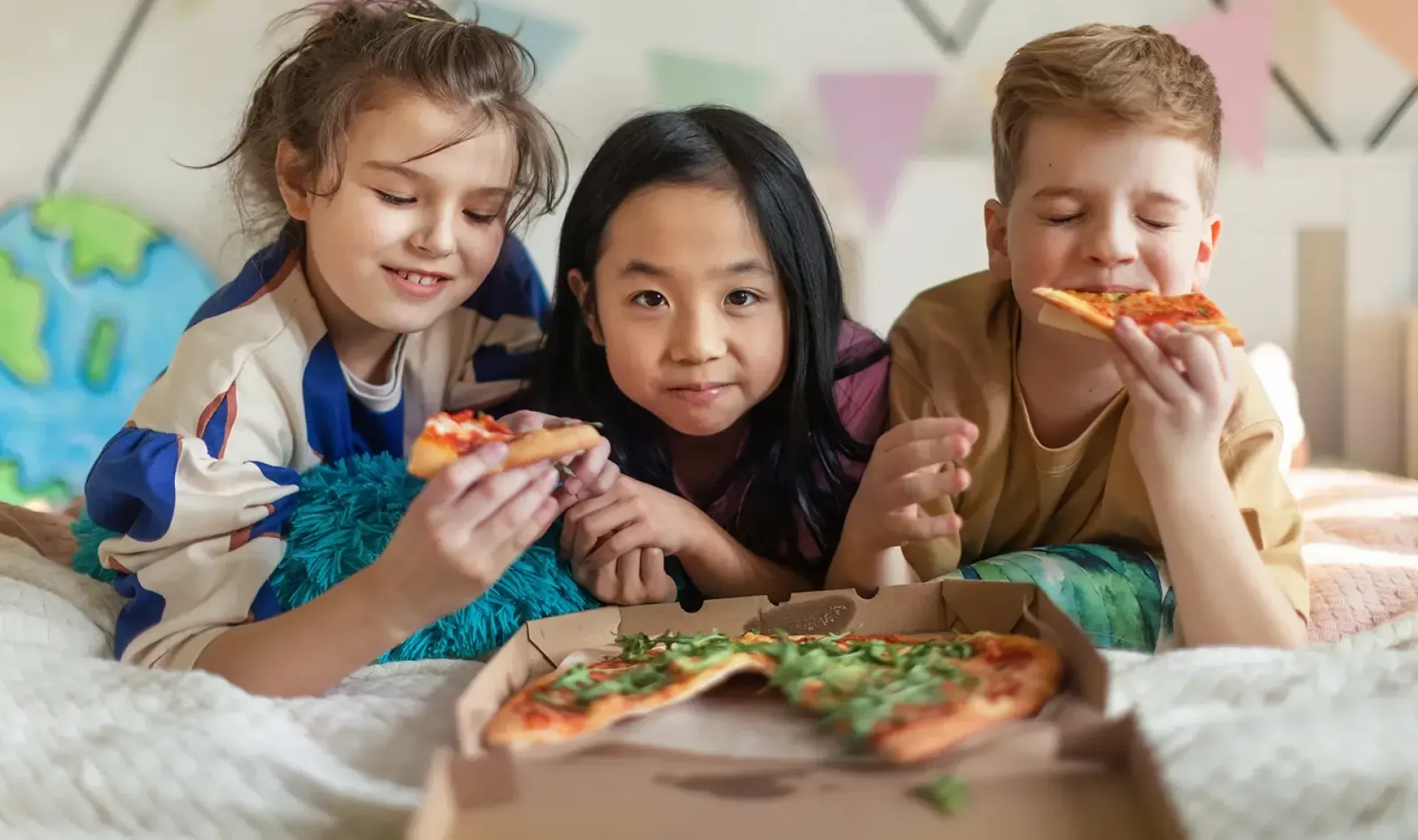 Three kids laying on their stomachs and eating pizza.