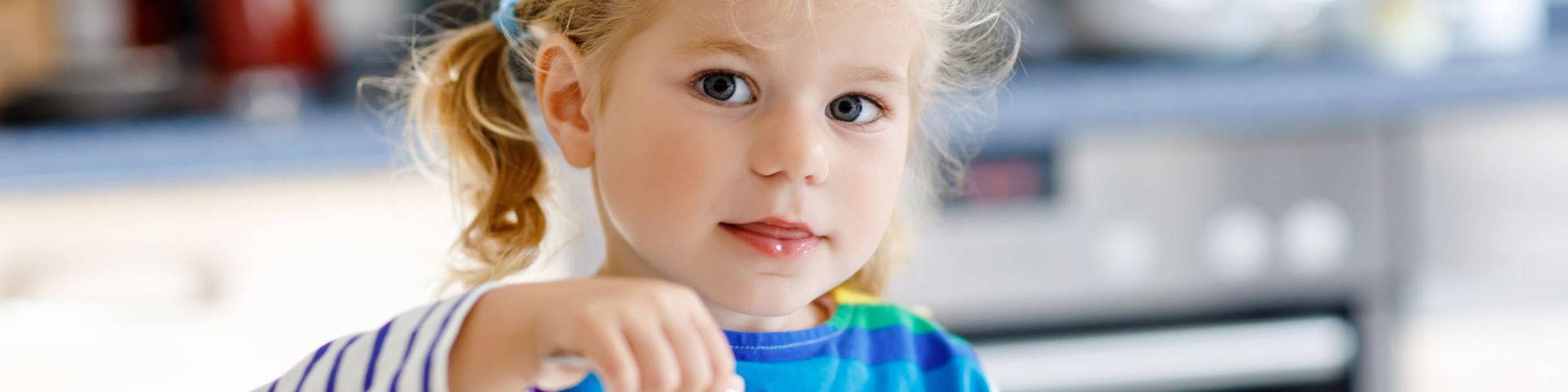 Blond child with pigtails wearing a rainbow striped shirt, eating from a bowl while seated at a table.