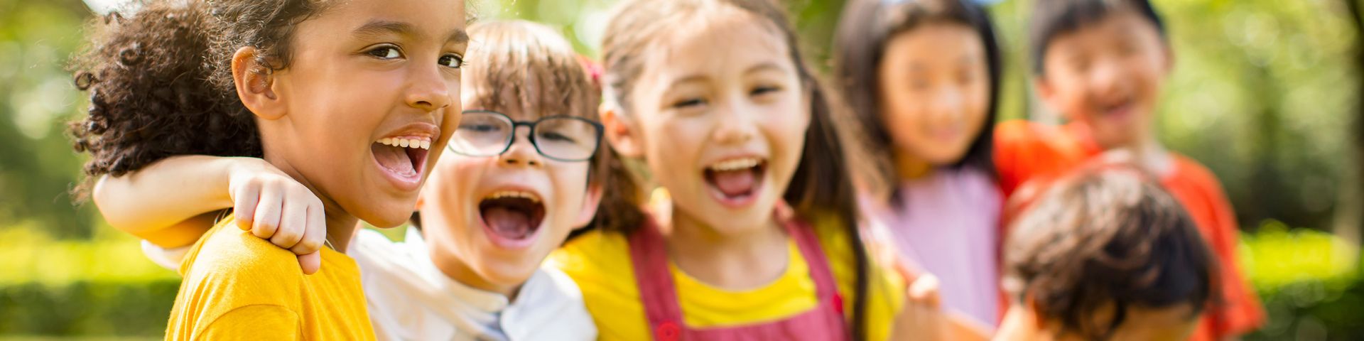 Kids with a variety of skin tones wearing colorful clothing outdoors in the sunshine, standing in a line with arms wrapped around each other's shoulders, laughing.