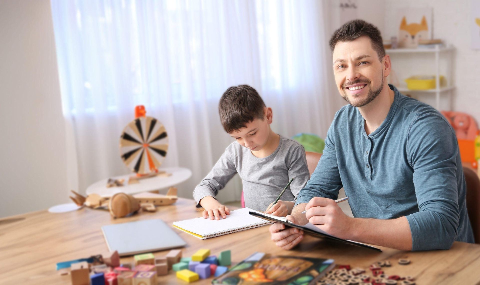 A plain clothed man with clipboard seated near child writing in notebook.
