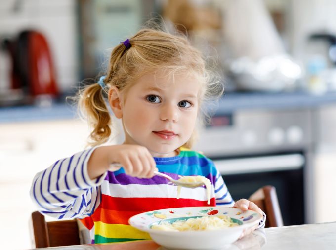 Blond child with pigtails wearing a rainbow striped shirt, eating from a bowl while seated at a table.