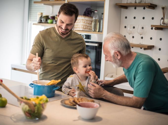 Middle aged adult male and older adult male prepare food in home kitchen while interacting with preschool boy seated at counter.