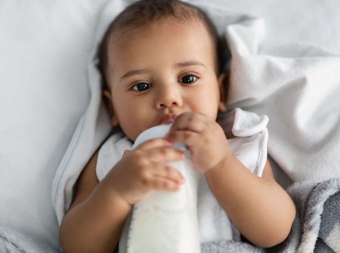 Infant with brown skin and dark hair laying in white bedding, drinking milk from a bottle.