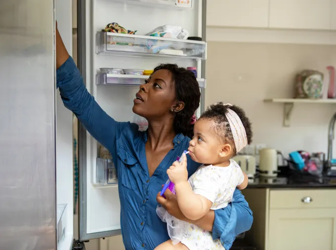 Black mother with infant daughter at open fridge.