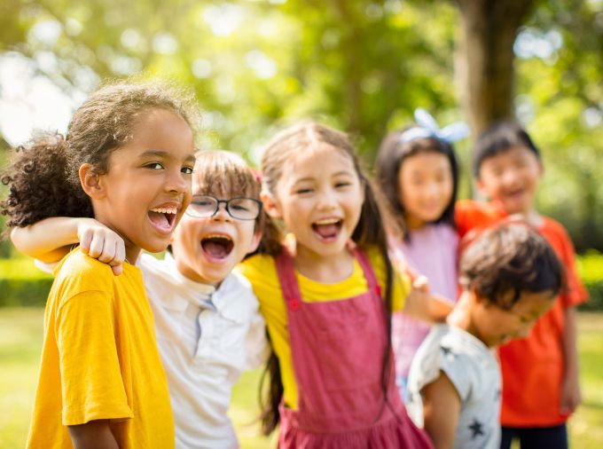 Kids with a variety of skin tones wearing colorful clothing outdoors in the sunshine, standing in a line with arms wrapped around each other's shoulders, laughing.