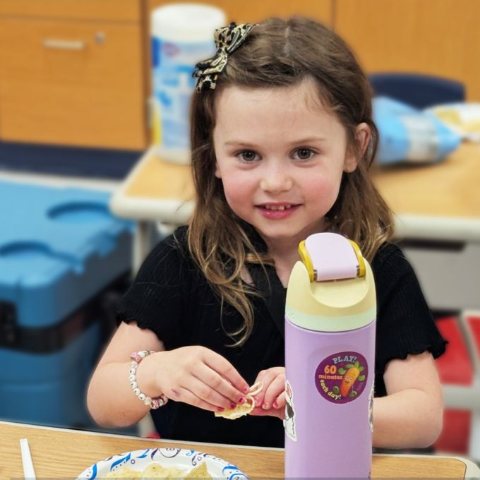 Child seated at classroom table with water bottle, eating.