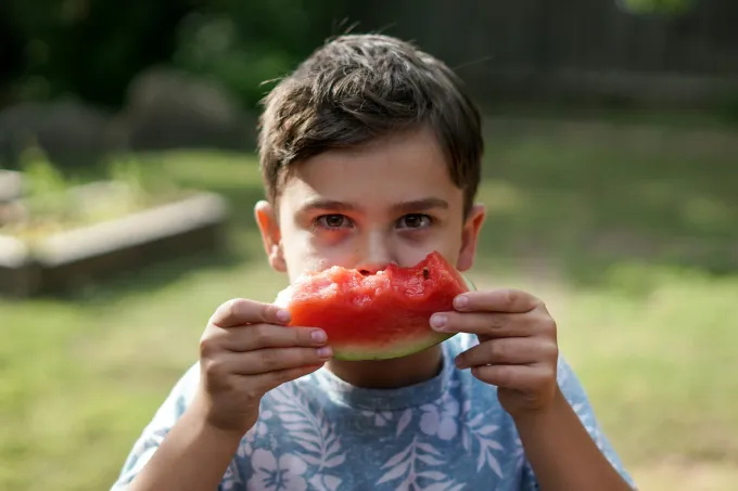 Boy, outside, holds partially eaten watermelon slice in front of face.