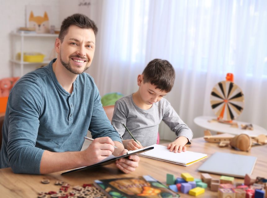 A plain clothed man with clipboard seated near child writing in notebook.
